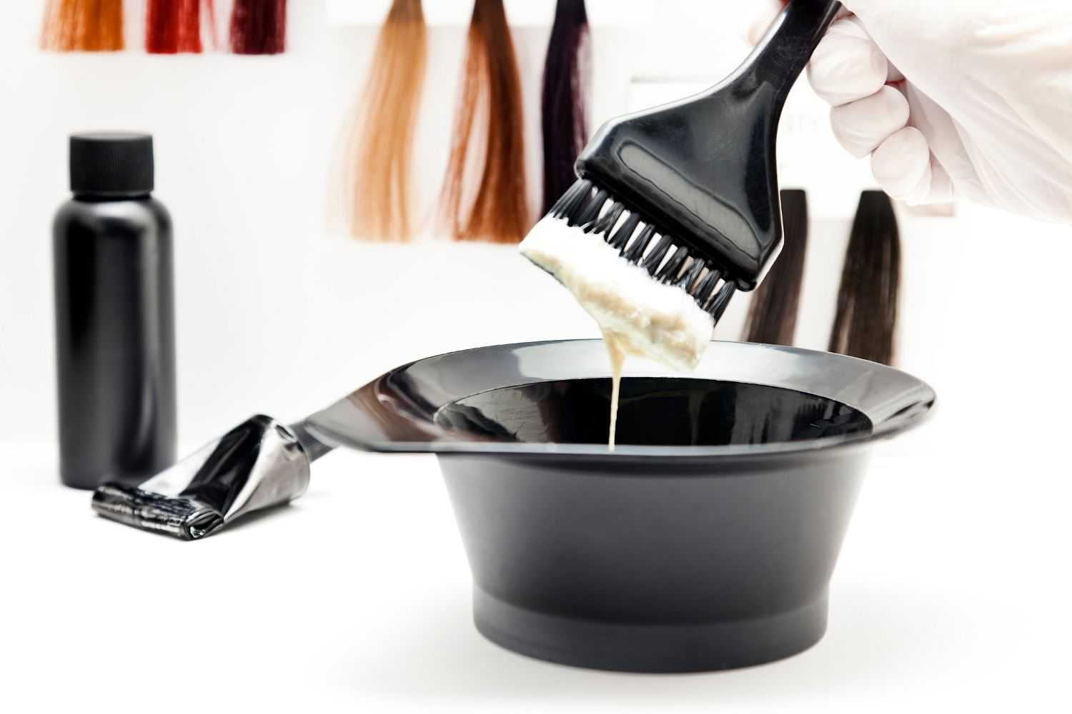 Hair dye being mixed in a black bowl with a brush, surrounded by color samples and a dye bottle.